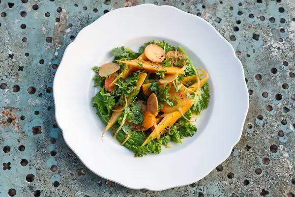 Salade colorée de carottes confites, parsemée d'herbes fraîches et de fines lamelles de radis, sur une assiette blanche.